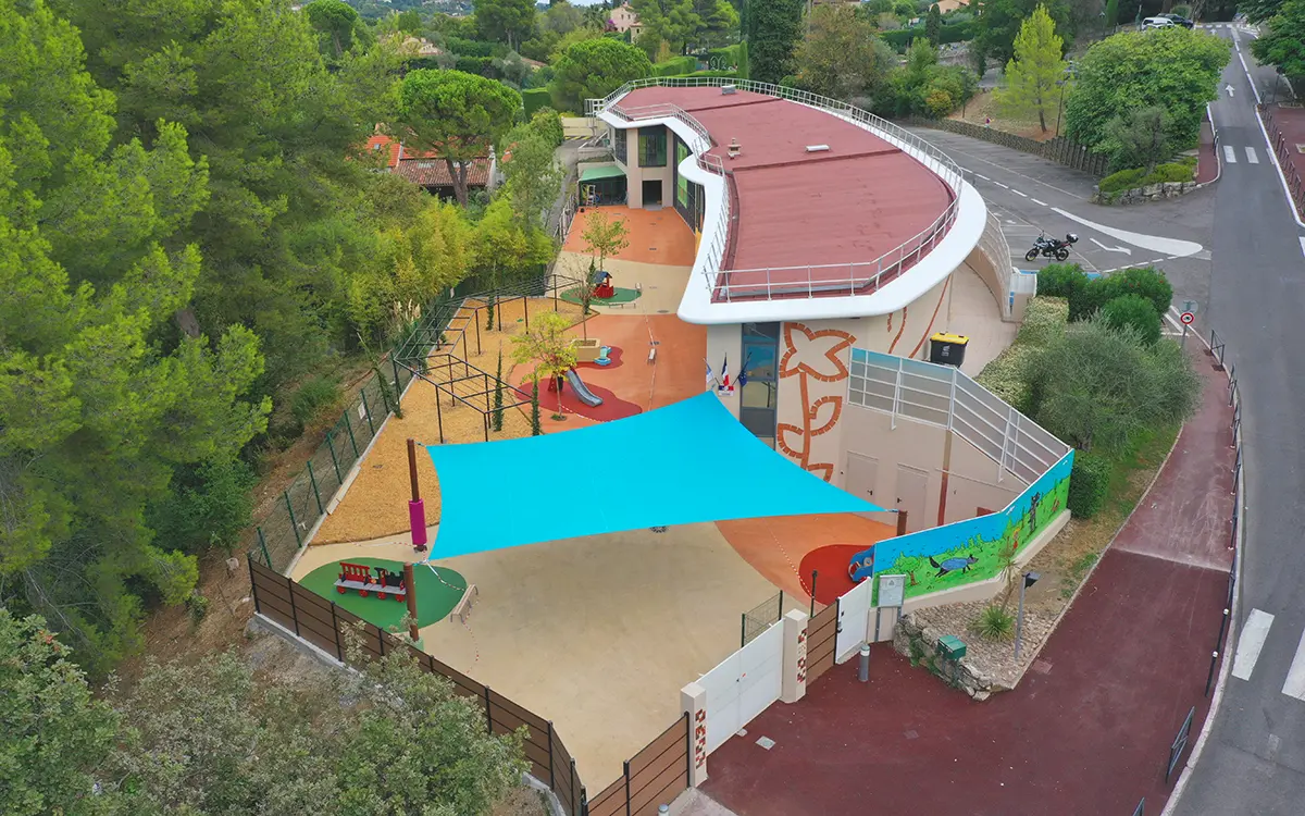 reinventer-et-re-enchanter-nos-ecoles_10 Une aire de jeux moderne avec toboggans, entourée d'arbres, près d'un bâtiment incurvé et routes. Couleurs vives et structures ludiques pour enfants.
