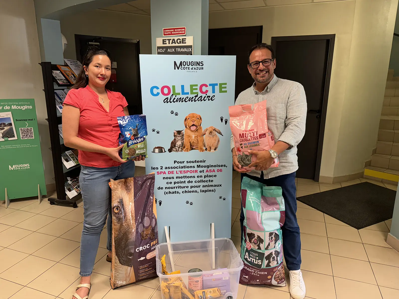 Mieux vivre avec nos animaux de compagnie_4 Deux personnes dans un hall participent à une collecte alimentaire pour animaux à Mougins, Côte d'Azur, avec des sacs de nourriture pour animaux.
