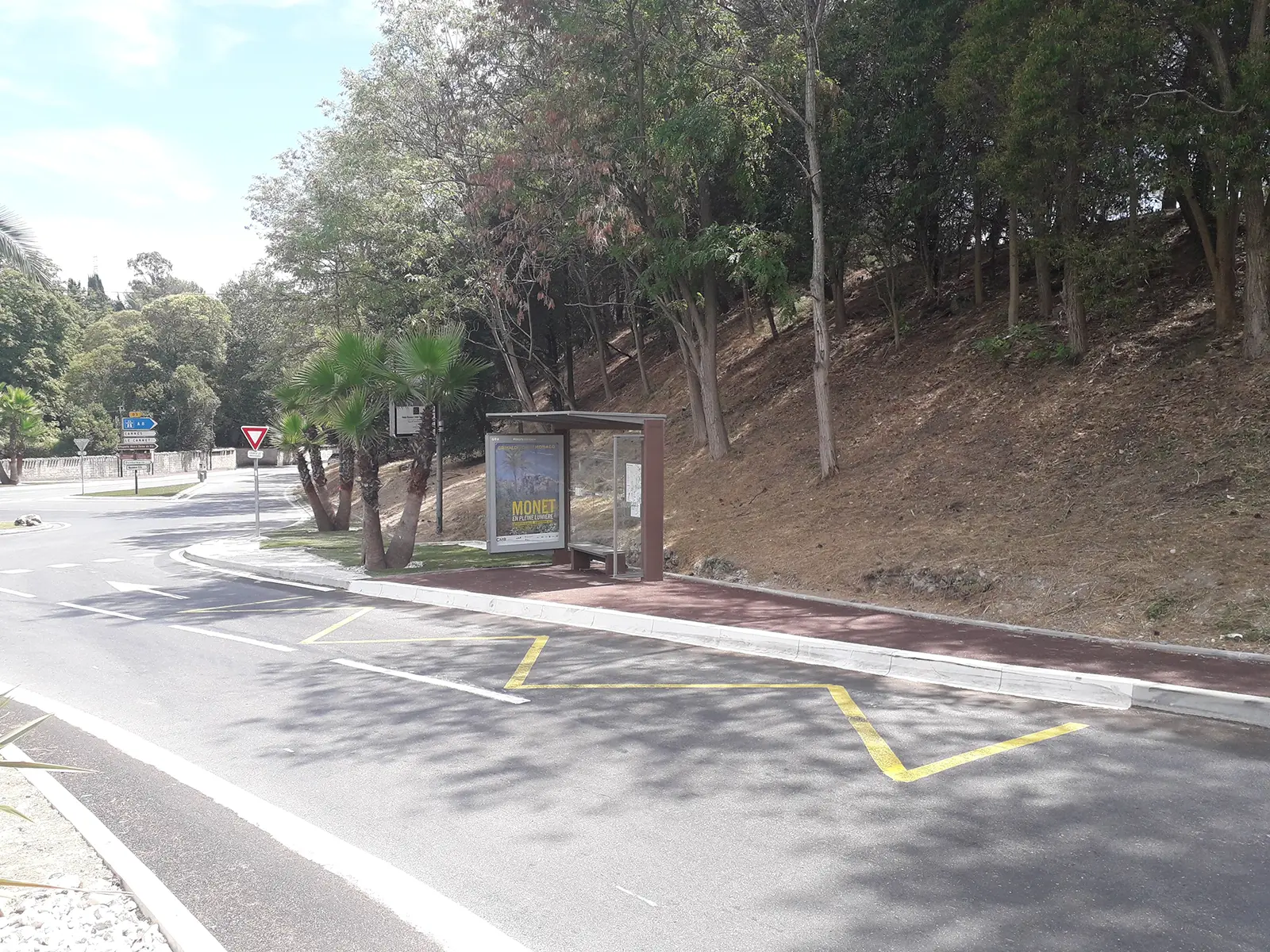 mise en accessibilité_2 Un abri de bus vide à côté d'une route bordée d'arbres, avec un panneau publicitaire et un panneau de signalisation routière.